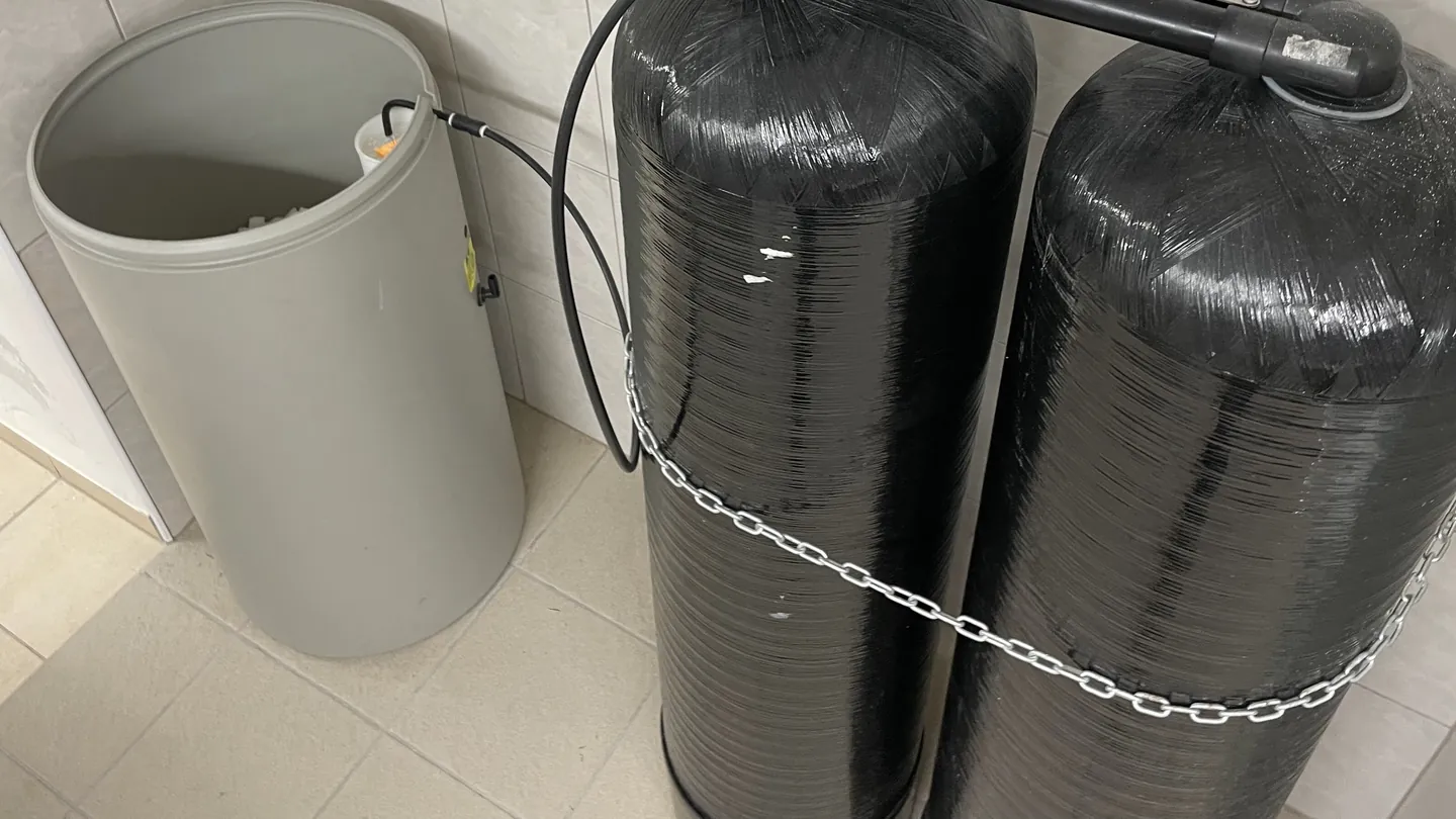 Two black resin tanks chained to the wall beside a gray brine tank on a tiled floor